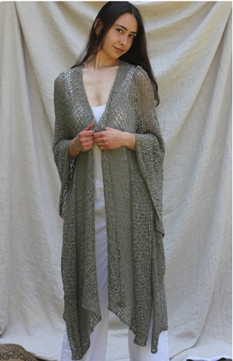 Person standing in front of draped fabric wearing a long, open-front olive knit cardigan over a white outfit.